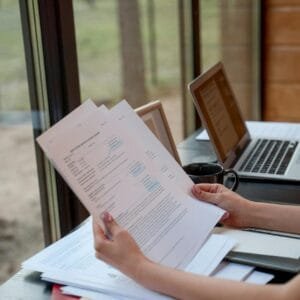 Person holding and reviewing documents near window with laptops on a desk.